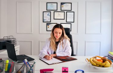 Nutritionist sits at a modern office desk writing, with fruits and diplomas displayed, emphasizing health and professionalism.